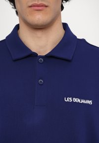 Gros plan d'une personne portant un polo bleu marine à col avec deux boutons et "LES BENJAMINS" brodé sur la poitrine.