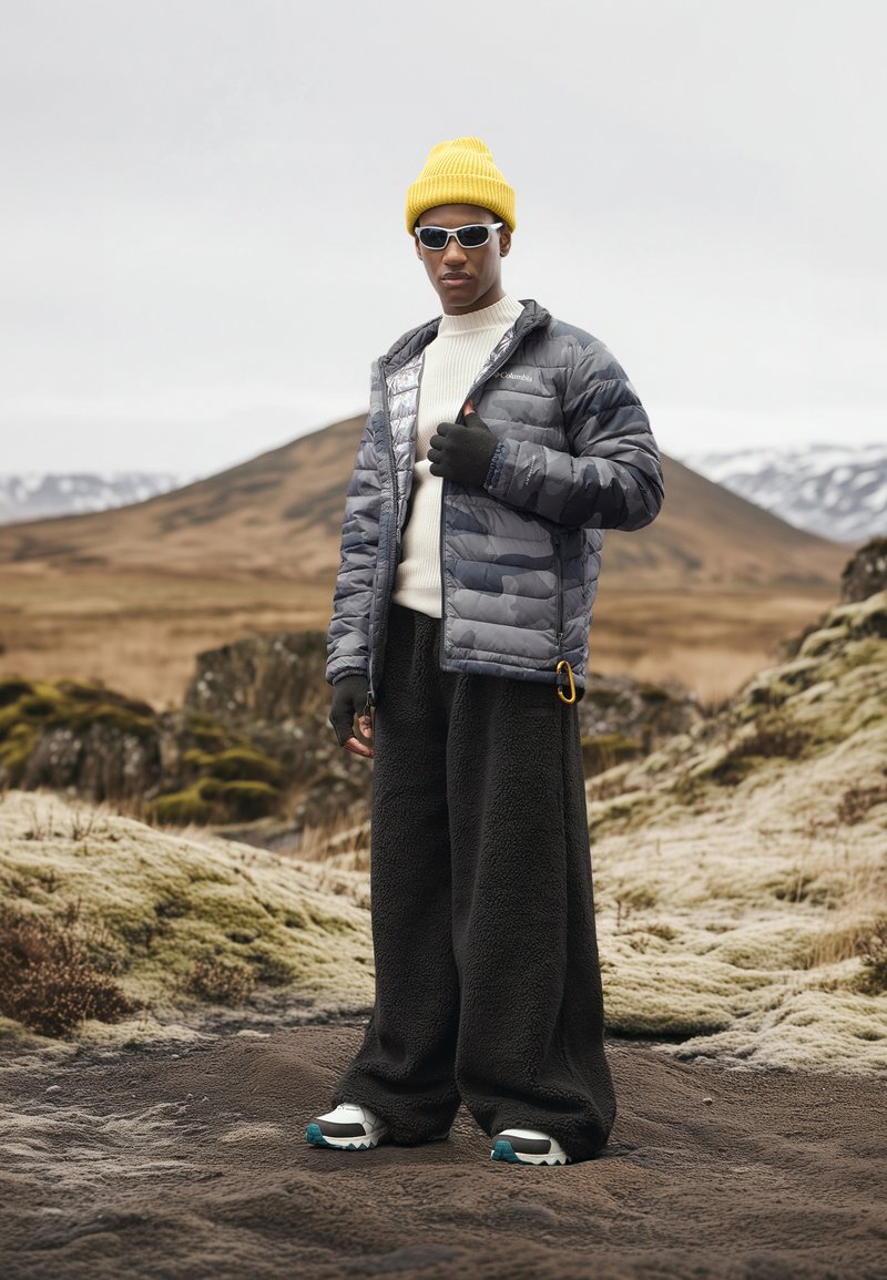 Chaqueta gris acolchada con un acabado brillante, suéter blanco, pantalones de forro polar negros, gorro de punto amarillo, gafas de sol y zapatos de senderismo gris y azul.