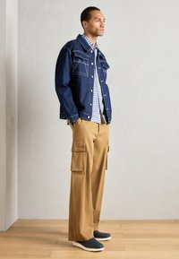 Veste en denim au design structuré, avec deux poches poitrine, associée à une chemise à carreaux bleus et un pantalon cargo beige, accentuée par des chaussures à enfiler.