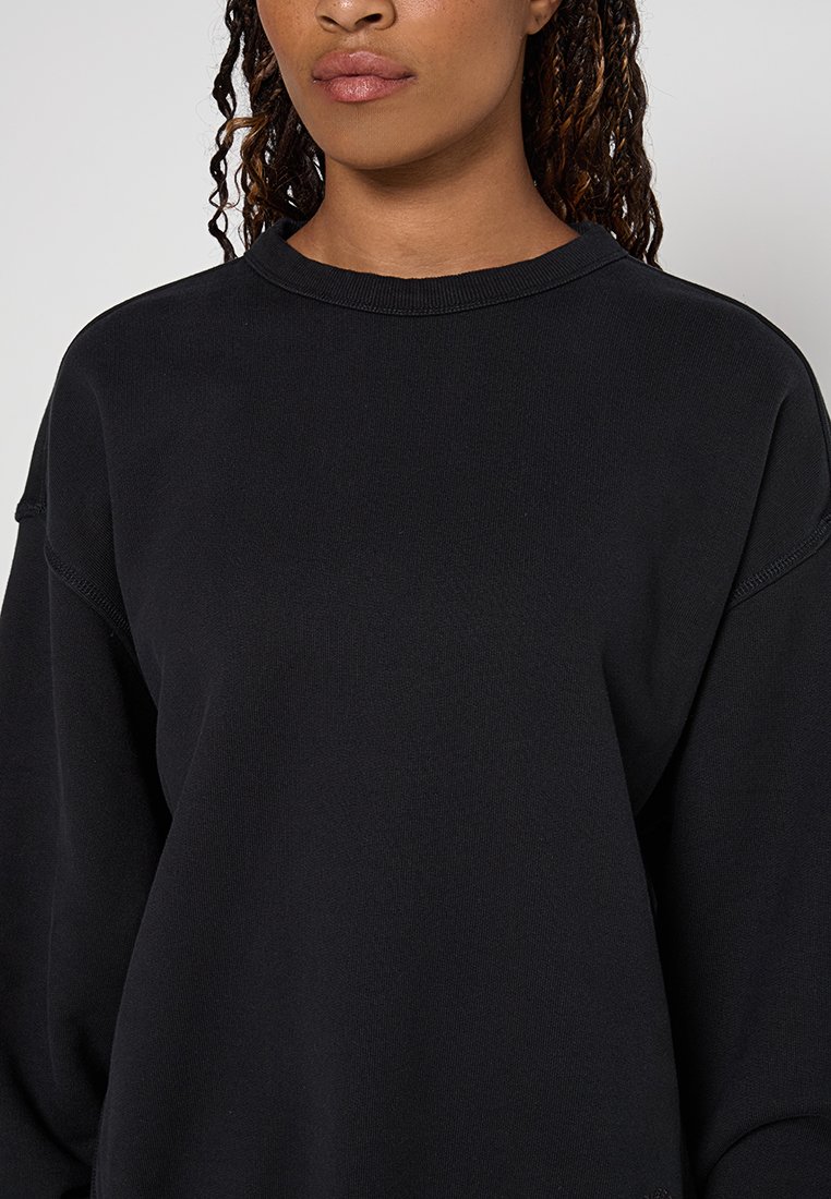 Donna con pelle di tonalità media-scura che indossa un semplice pullover nero a girocollo, mostrata dalle spalle fino a metà torace su uno sfondo neutro.