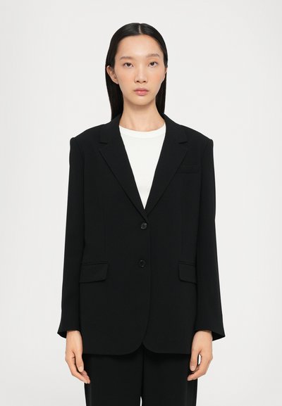 Blazer nero con spalle strutturate, doppio bottone, due tasche frontali e revers a scannatura, indossato su una camicia bianca. Tessuto liscio.