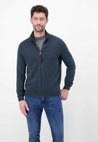 Donkergroene sweater met ritssluiting, textuurgebreid patroon, geribbelde manchetten en een hoge kraag, gedragen over een grijs shirt en gecombineerd met een blauwe spijkerbroek.