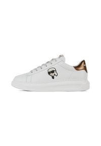 Sneaker in pelle bianca con un accento in pelle lucida marrone sul tallone, lacci argentati e un logo di carattere nero sul lato. Suola arrotondata e texturizzata.