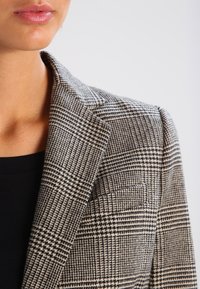 Blazer med svart och krämfärgad rutmönster, med slagkrage och en enkel bröstficka. Tillverkad av texturerad tyg.