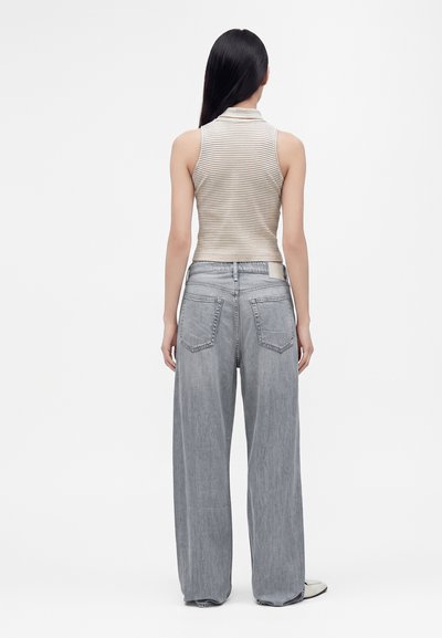 Femme aux longs cheveux foncés, portant un haut beige rayé sans manches et un jean ample gris clair, debout de dos sur un fond blanc uni.