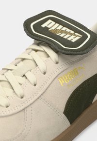 Sapatilhas de camurça bege com detalhes em camurça verde e marcação em dourado. Apresenta uma sola grossa e atacadores texturizados. Etiqueta com o logótipo em destaque.