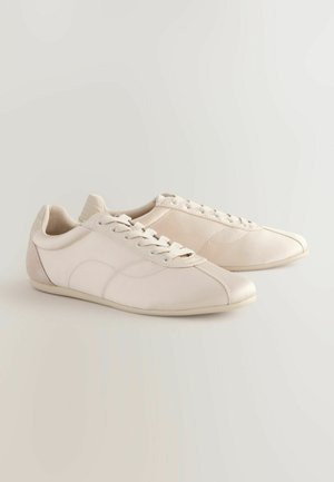 Pasiltai kreminiai sportiniai batai, pagaminti iš sintetinių medžiagų, turintys lygų paviršių, su raištelių užsegimu ir subtiliais siuvinėjimų raštais.