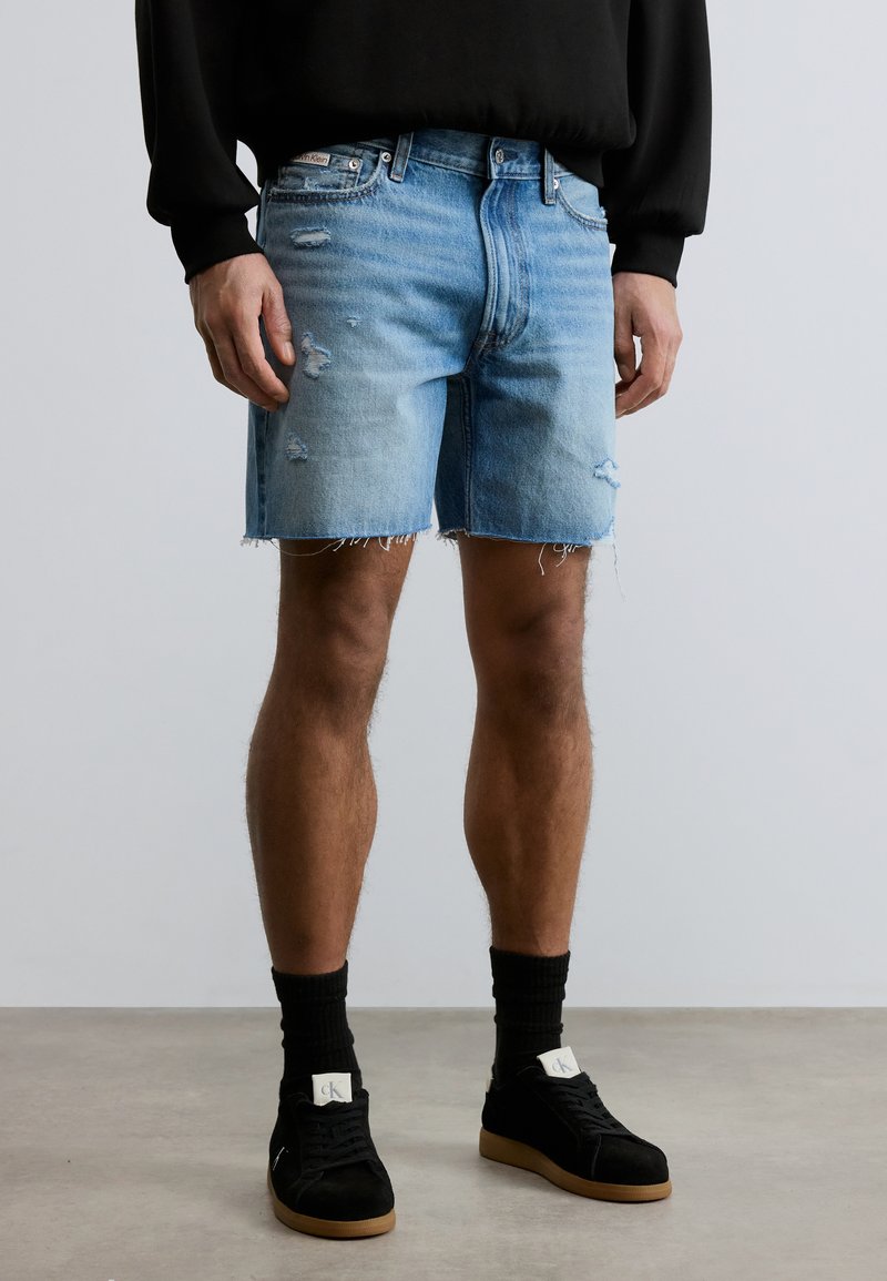 Άνδρας με ανοιχτό μπλε τζιν σορτς με φθορές, μαύρο μακρυμάνικο πουκάμισο, μαύρες κάλτσες και μαύρα αθλητικά παπούτσια με καφέ σόλες, σε απλό φόντο.