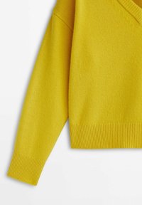 Manche et ourlet d'un pull jaune tricoté avec des poignets et une ceinture côtelés sur un fond blanc.