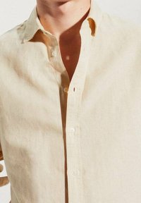 Chemise beige clair boutonnée, portée ouverte au col, laissant voir une partie du cou et du haut de la poitrine.