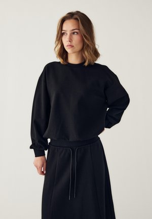 Ung kvinde med skulderlangt hår iført en sort langærmet sweatshirt og matchende sort nederdel med snørebånd, ser til siden.