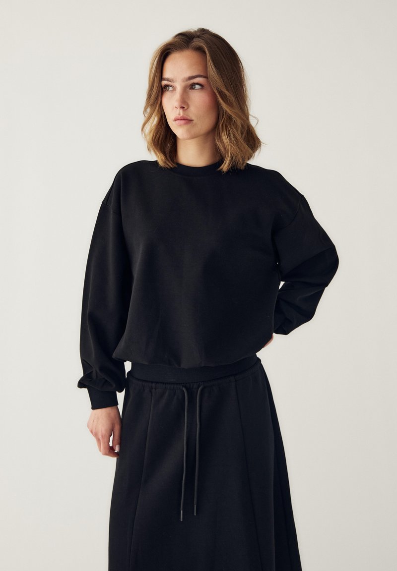Jeune femme aux cheveux mi-longs portant un sweat-shirt noir à manches longues et une jupe noire assortie avec cordon, regardant sur le côté.