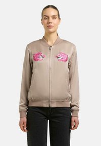 Kobieta ubrana w beżową satynową kurtkę bomber z różowymi, haftowanymi smokami trzymająca róże na piersi, w połączeniu z czarnymi jeansami.