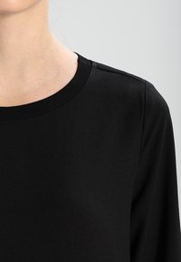 Musta pyöreäkauluksinen neulepaita, joka on valmistettu pehmeästä kankaasta. Siinä on ribbireunat kauluksessa ja pitkissä hihoissa, sileä pinta ja minimalistic design.