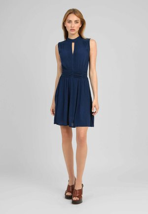 Femme portant une robe bleu marine sans manches avec un décolleté en forme de goutte et des talons compensés marron ouverts à l’avant, debout devant un fond gris uni.