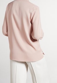 Helle rosa Bluse mit langen Ärmeln, glatter Textur, rundem Ausschnitt und seitlichen Schlitzen, kombiniert mit weißen weiten Hosen.