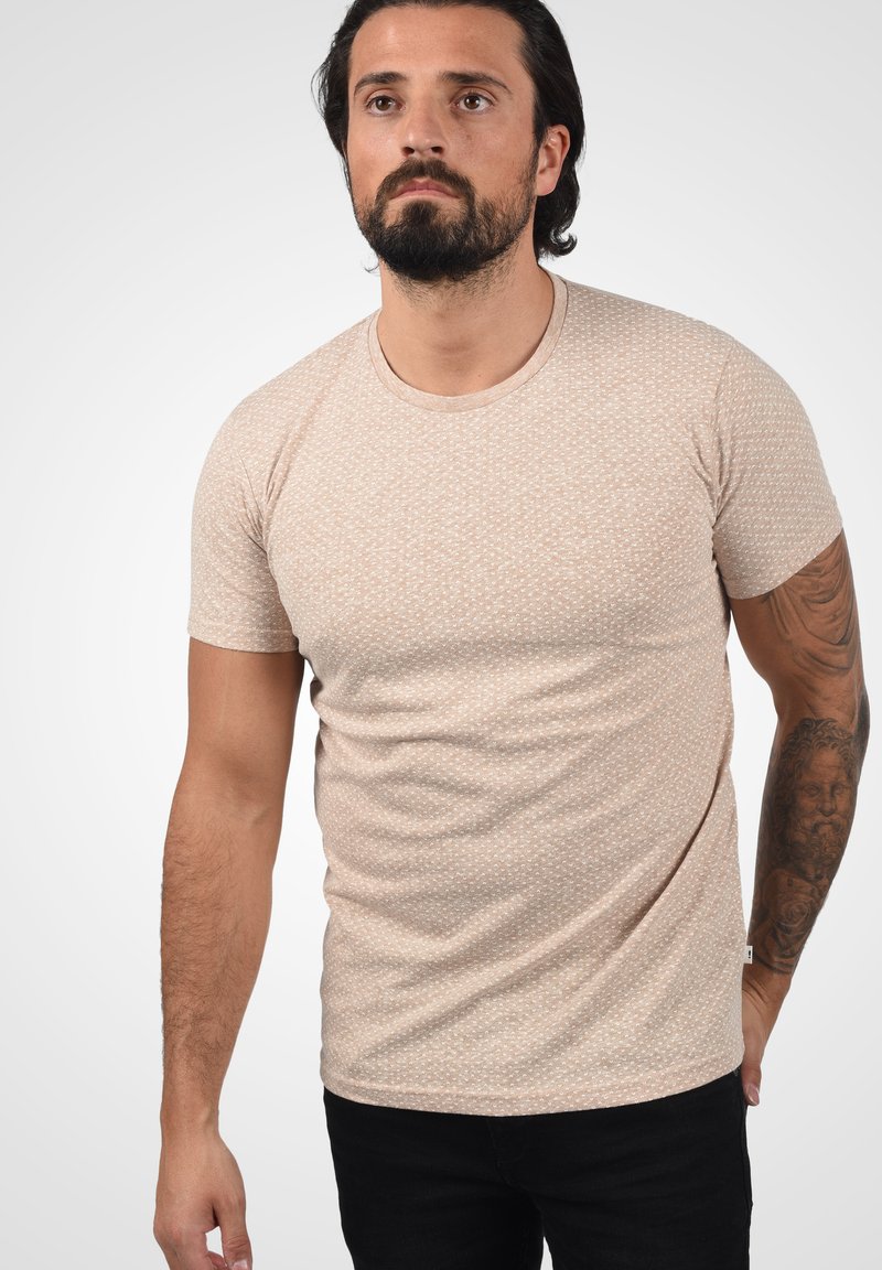 Camiseta beige de manga corta con un patrón sutil, cuello redondo y una forma ajustada, elaborada en una tela suave. Combinada con jeans negros.