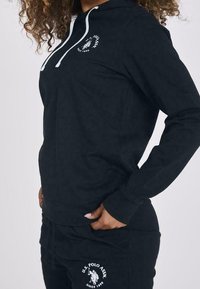 Felpa nera con cappuccio e lacci bianchi, caratterizzata da un logo sul petto e pantaloni neri abbinati con un logo ricamato in bianco.
