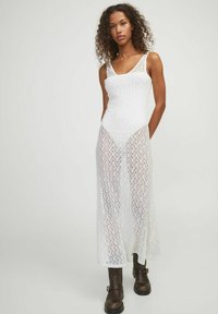 Vestido blanco hasta larodilla con un diseño de punto texturizado y abierto, con tirantes anchos y un corsé ajustado, combinado con botas hasta el tobillo marrones.