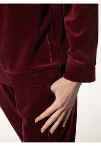 Bordeaux fluwelen trainingspak met een zachte textuur, voorzien van een zijzak en decoratieve stiksels op de naad, die een modern ontwerp tentoonstelt.