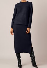 Granatowy dzianinowy komplet dwuczęściowy o prążkowanej fakturze, z długimi rękawami i spódnicą do kolan z bocznymi rozcięciami. Czarne kozaki za kolano.