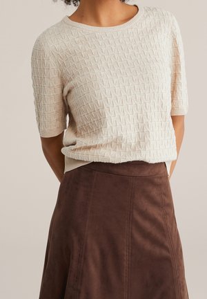 Femme portant un haut beige texturé à manches courtes, rentré dans une jupe marron en daim taille haute, sur un fond uni.
