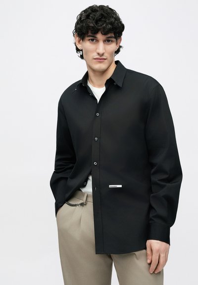 Chemise noire à manches longues avec col boutonné et une étiquette discrète, en tissu lisse, portée sur un t-shirt blanc.