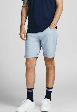 Mand iført lyseblå shorts med opsmøg, marineblå skjorte, sorte stribede sokker, hvide sneakers, tatoveringer synlige på venstre arm.