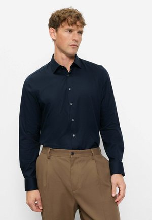 Mann mit lockigem Haar, der ein navyblaues, langärmliges Hemd mit Knöpfen trägt, in die braune Hose gesteckt, und vor einem weißen Hintergrund steht.
