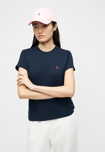 Νεαρή γυναίκα με ροζ καπέλο μπέιζμπολ, μπλε σκούρο μπλουζάκι με κόκκινο λογότυπο και λευκό παντελόνι, στέκεται με τα χέρια σταυρωμένα μπροστά σε απλό φόντο.