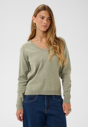 Femme aux longs cheveux blonds portant un pull en maille vert à col en V et un jean bleu, debout devant un fond clair uni.