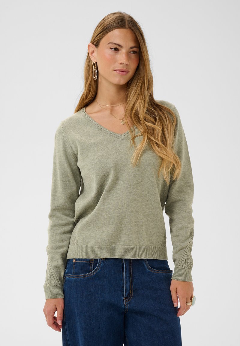 Femme aux longs cheveux blonds portant un pull en maille vert à col en V et un jean bleu, debout devant un fond clair uni.