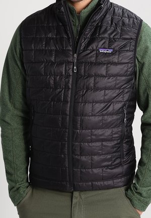 Mand iført en sort quiltet Patagonia-vest over en grøn langærmet trøje med lynlås, med hænderne i lommerne på olivengrønne bukser.