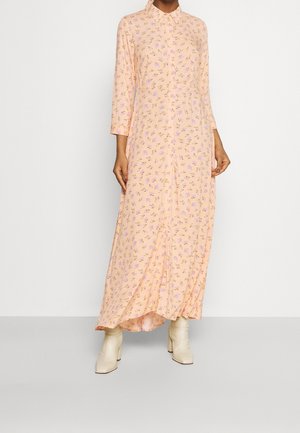 Femme portant une longue robe fleurie pêche avec des boutons, des manches trois-quarts et des bottines crème sur un fond uni.