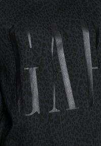 Sweat-shirt noir avec une texture imprimé léopard et un logo "GAP" audacieux et réfléchissant au centre. Tissu lisse avec un col rond.