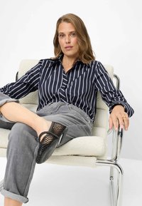 Marineblauw gestreept overhemd met knopen, grijze opgerolde spijkerbroek en zwarte gevlochten schoenen. Model zittend op een moderne chromen stoel tegen een witte achtergrond.