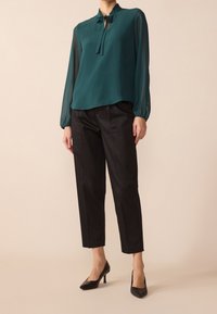 Blouse teal à col montant, avec un détail à nouer et des manches longues transparentes, associée à un pantalon noir à jambes droites et des escarpins noirs.