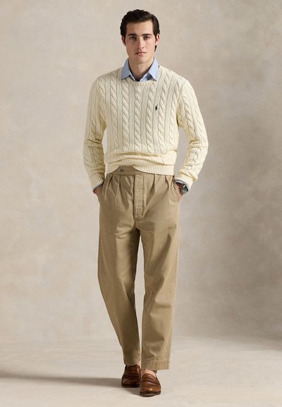 Jeune homme debout, les mains dans les poches, portant un pull torsadé crème, une chemise bleu clair, un pantalon plissé beige et des mocassins marron.