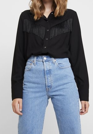 Femme portant un haut à manches longues noir avec des détails à franges sur la poitrine et un jean bleu clair taille haute.