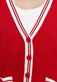 Cárdigan rojo con ribete a rayas en blanco y rojo, abrochado con cuatro botones rojos sobre una camiseta blanca con canalé.