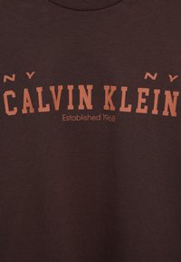 Tamnosmeđa majica kratkih rukava s natpisom "CALVIN KLEIN" u podebljanom narančastom slovu, a ispod manji narančasti font s natpisom "Established 1968".