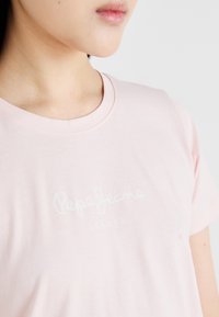 T-shirt de algodão rosa claro com mangas curtas, apresentando um logo branco "Pepe Jeans" e o texto "LONDON" ao centro. Textura suave.