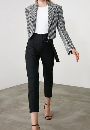 Frau trägt schwarze, gekürzte Hose mit Gürtel, weißes Top und schwarz-weißes Hahnentritt-Cropped-Blazer, geht auf einem Betonboden.