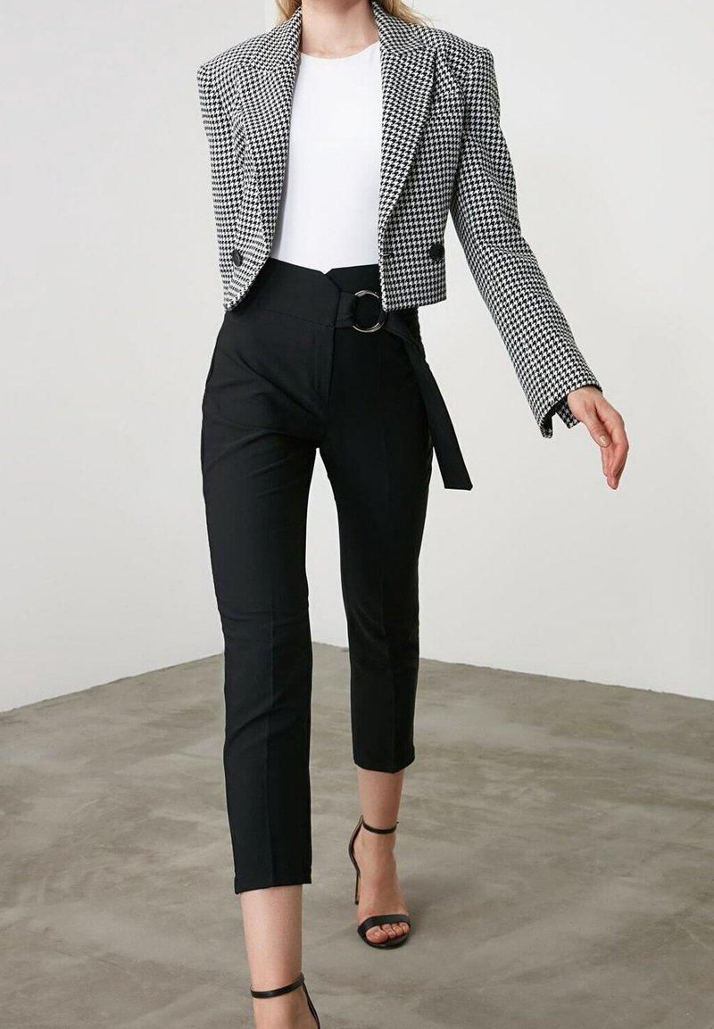 Femme portant un pantalon noir raccourci avec ceinture, un haut blanc et un blazer court pied-de-poule noir et blanc, marchant sur un sol en béton.