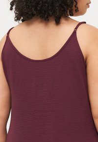 Maroon tank top with thin, adjustable straps, a smooth, textured fabric, and a rounded neckline, showing the back design.