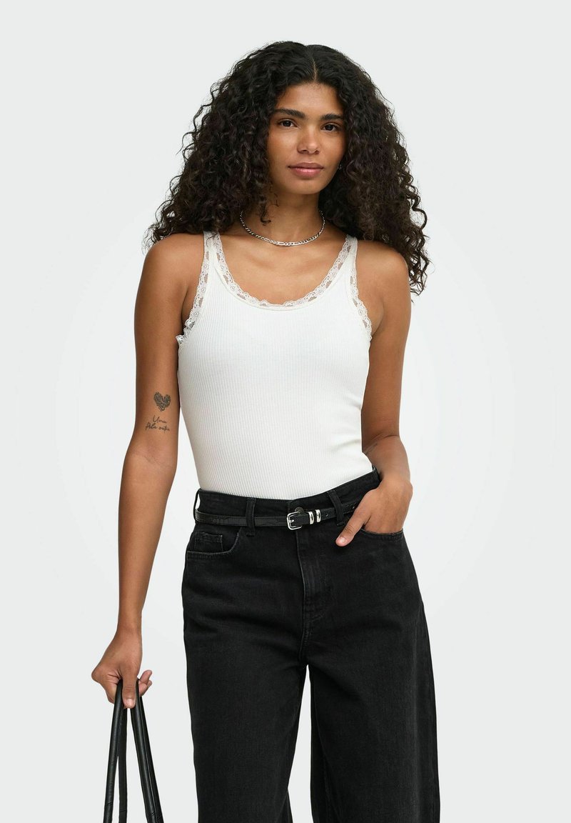 Femme aux cheveux bouclés portant un débardeur blanc bordé de dentelle et un jean noir taille haute, tenant un sac à main noir sur un fond uni.