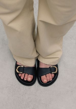 Sandales glissantes en cuir noir avec un détail de boucle dorée. Les pieds portant les sandales sont posés sur un sol gris clair, avec un pantalon beige couvrant légèrement les chevilles.