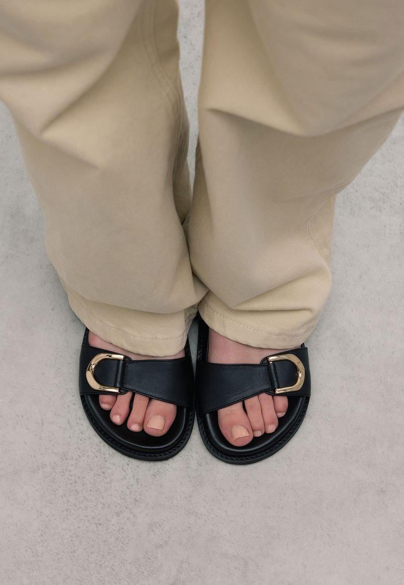 Sandales glissantes en cuir noir avec un détail de boucle dorée. Les pieds portant les sandales sont posés sur un sol gris clair, avec un pantalon beige couvrant légèrement les chevilles.