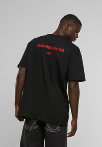 Black cotton t-shirt with short sleeves, featuring red text "Osaka • Paris • New York" on the back. Loose fit with a round neckline.
