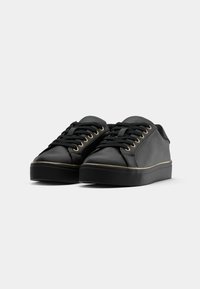 Sneakers neri con una finitura liscia e opaca, occhielli dorati, lacci piatti e una suola in gomma testurizzata. Presentano un dettaglio di rifinitura dorato a contrasto.